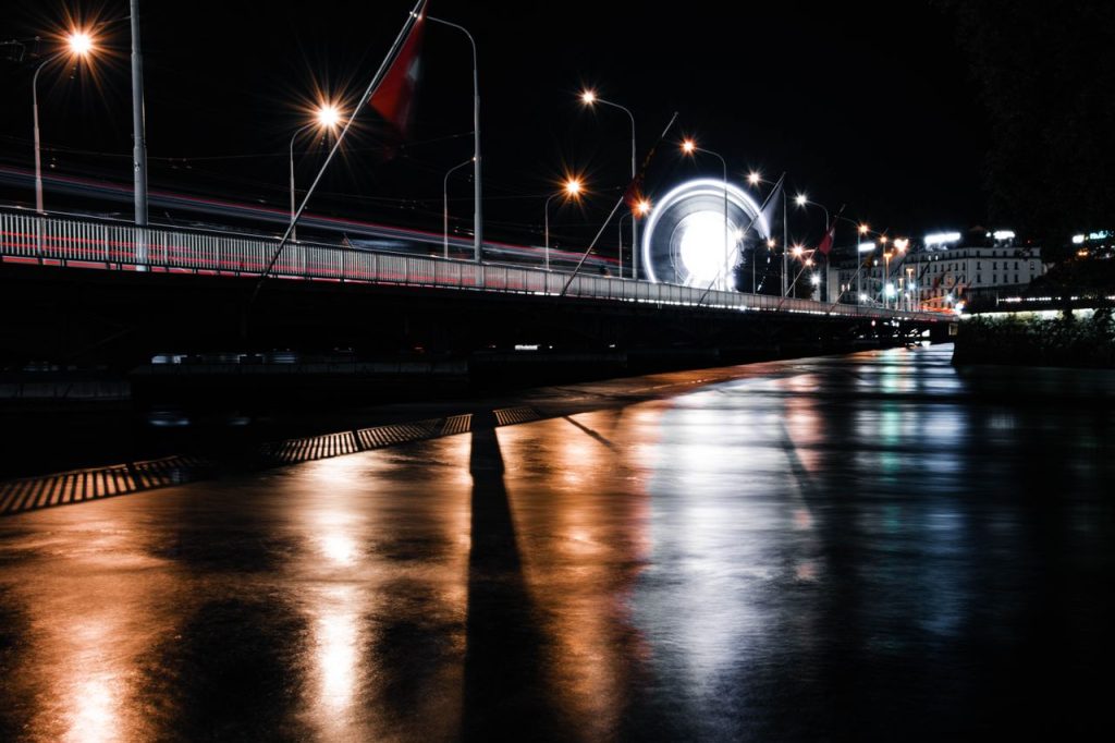 Stunning night view of Geneva bridge with illuminated ferris wheel, beautifully reflected in the wet pavement.